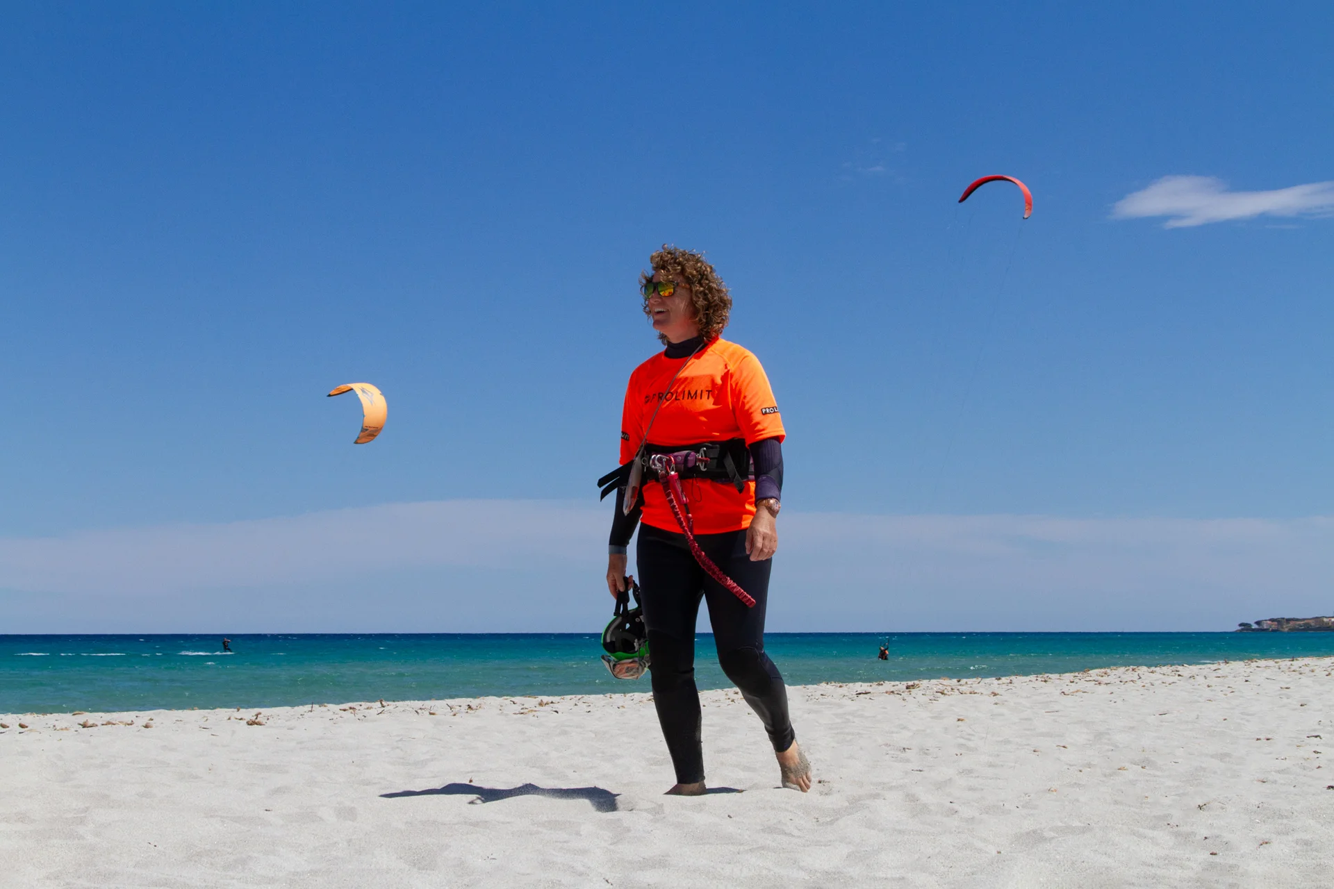 Istruttrice di kitesurf in divisa arancione sulla spiaggia, con kite in volo sullo sfondo.