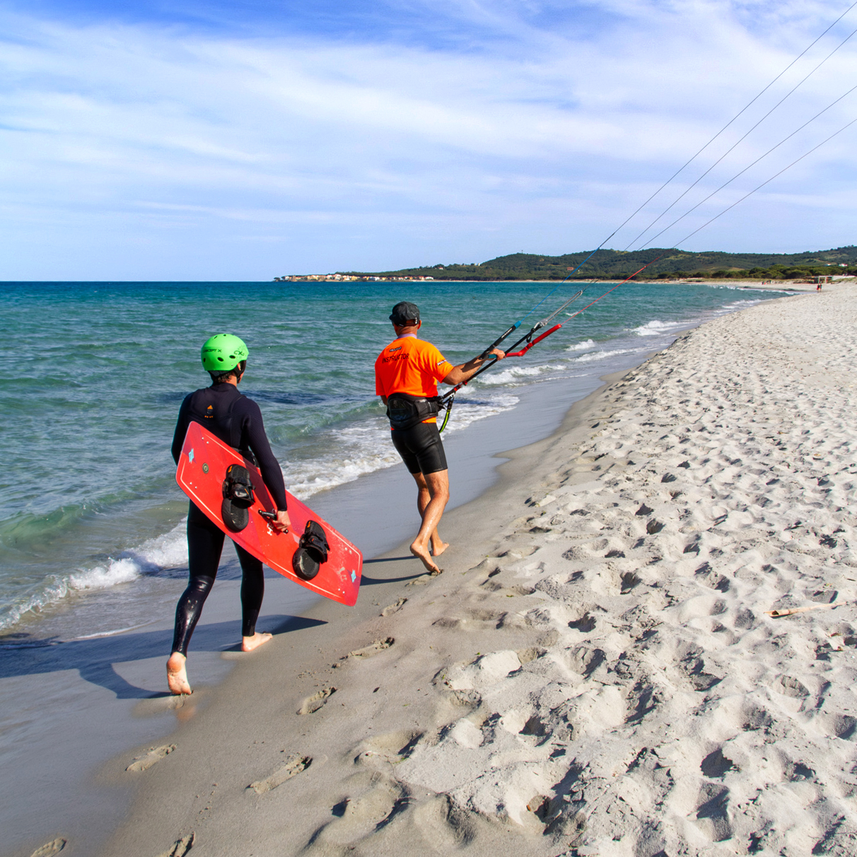 Zwei Kitesurfer gehen mit Board und Trapez am Strand entlang, bereit für eine Kitesurfstunde auf Sardinien